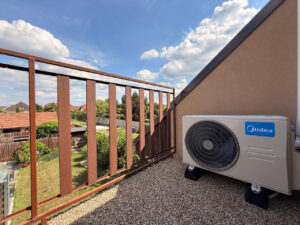 Venkovní jednotka Midea Multi-split 5,3 kW na antivibrační podstavce na balkóně v patře rodinného domu, letní obloha a zahrada v pozadí