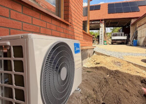 Venkovní jednotka Midea Single-split 3,5 kW upevněná na fasádní konzolové držáky na cihlové fasádě rodinného domu, v pozadí fotovoltaické panely na střeše